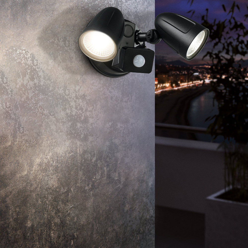 Black outdoor wall-mounted spotlight with a built-in motion sensor, shown mounted on a textured gray wall with a split view of its illuminated light beam and a nighttime cityscape background.