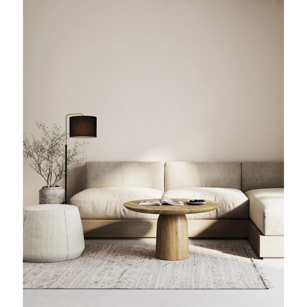 A modern living room setup featuring a beige sectional sofa, a round wooden coffee table, a textured pouf, a floor lamp, and a neutral rug against a warm beige wall.