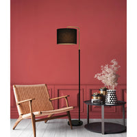 A modern black floor lamp with a curved gold-toned arm and black drum shade stands beside a woven wicker armchair and a small round side table with dried flowers, set against a terracotta red paneled wall.