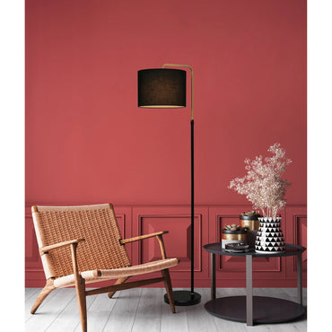 A modern black floor lamp with a curved gold-toned arm and black drum shade stands beside a woven wicker armchair and a small round side table with dried flowers, set against a terracotta red paneled wall.