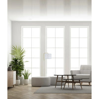 A bright, modern living room with three tall white-framed windows, a light gray armchair, a round ottoman, a low wooden coffee table, a floor lamp, and potted plants.