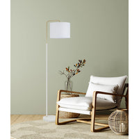 A modern floor lamp with a white drum shade and slim gold-toned metal frame stands beside a wooden armchair with white cushions in a styled room setting.