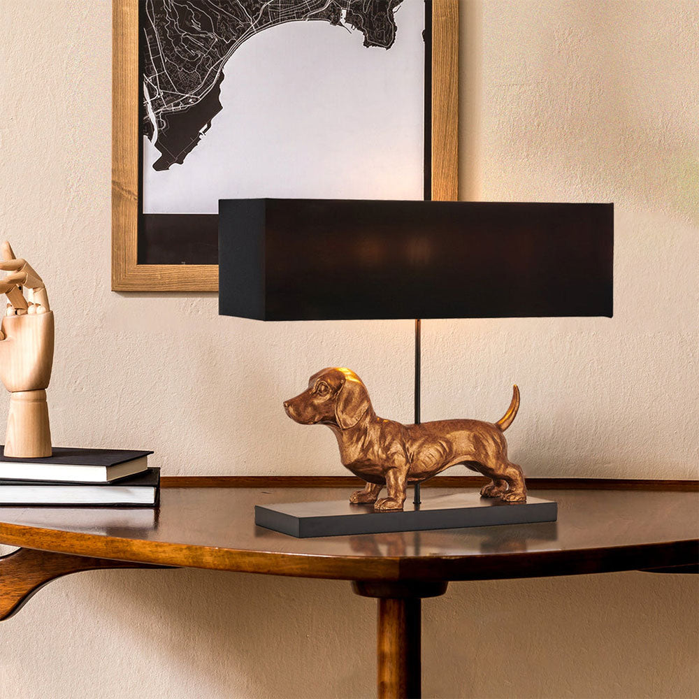 Dog-shaped table lamp on a wooden table with a black lampshade, framed artwork in the background.