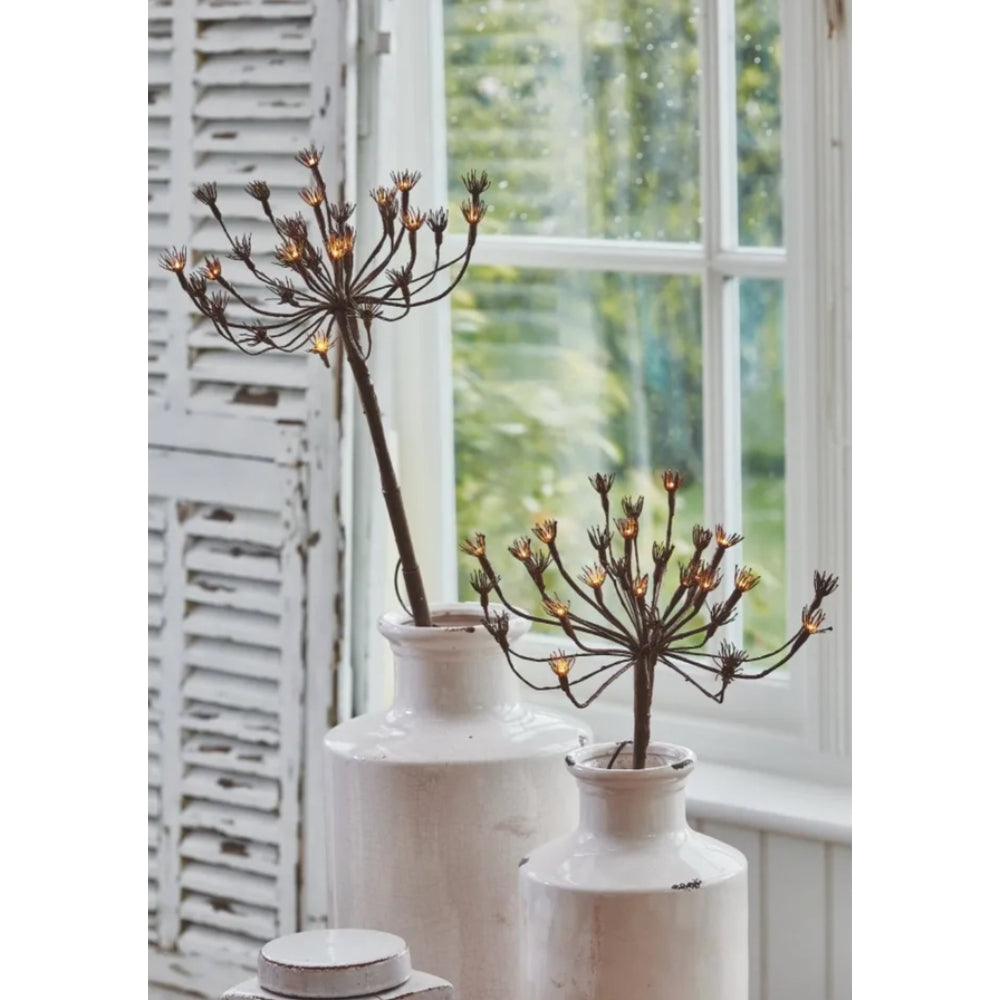 Decorative plant-like sculptures in white vases with a window and white shutters in the background.